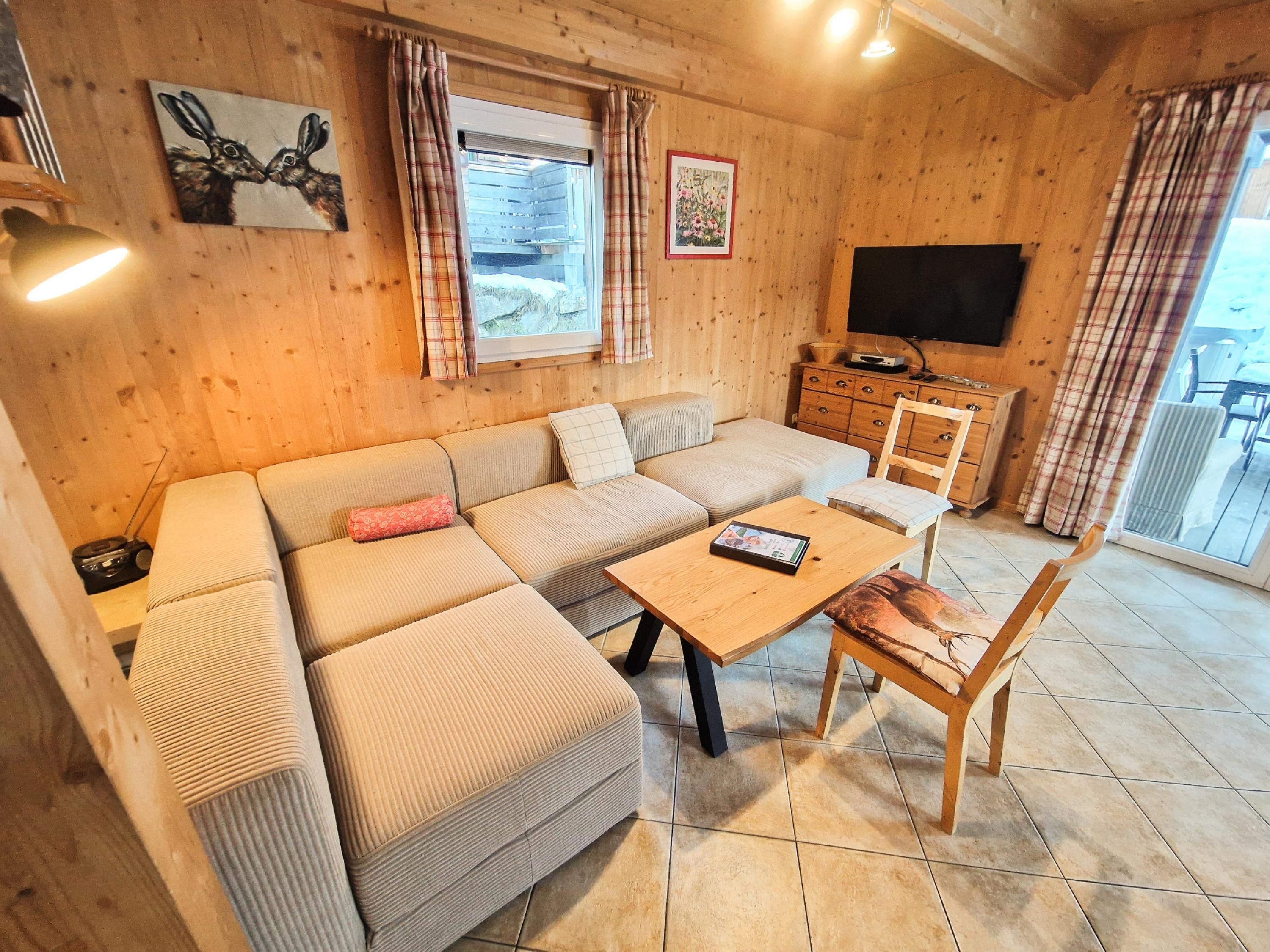 Ein gemütliches Blockhäuschen-Interieur mit einer Eckcouch, einem Holztisch, einem Stuhl, einem Fernseher und einem Fenster mit Vorhängen.