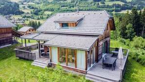 A wooden cabin with a glass-enclosed patio, surrounded by lush greenery and a scenic view of rolling hills.