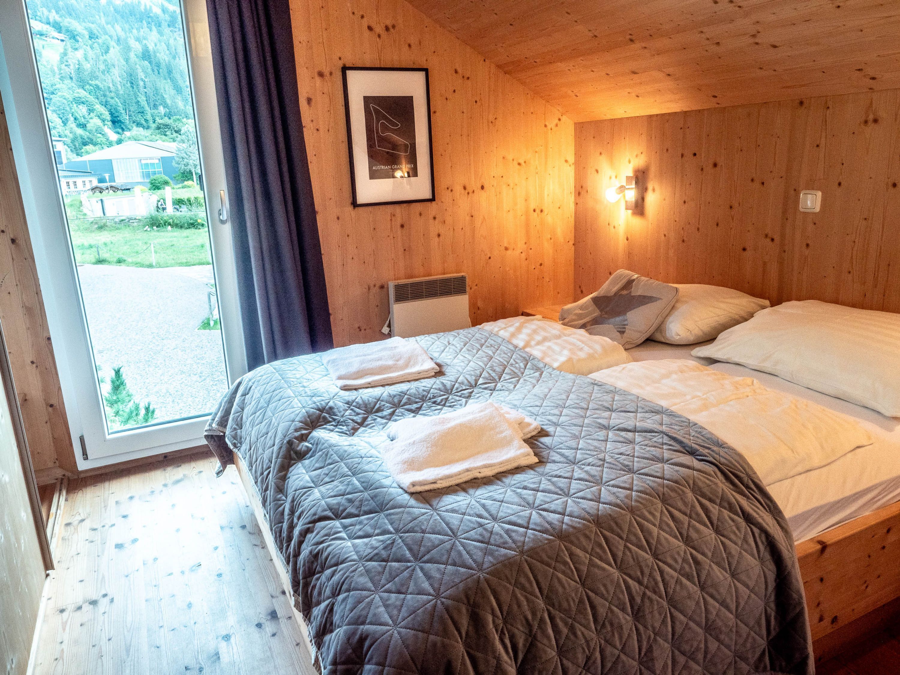 A wooden room with a bed, a framed picture on the wall, and a window with a view of greenery.