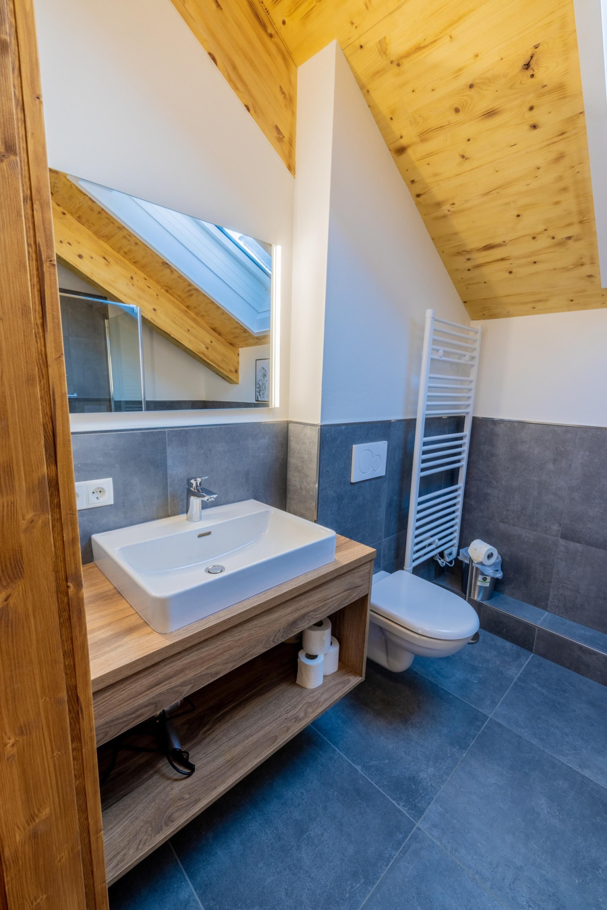 A modern bathroom with a wooden ceiling, a large window, a white sink, a toilet, and a towel rack.