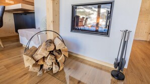 Ein Zimmer mit Holzboden, einem Fenster mit Blick auf eine verschneite Landschaft, einem Holzstapel und einem an der Wand angelehnten Besen.