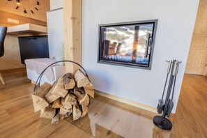 Una habitación con piso de madera, una ventana que ofrece vistas de un paisaje nevado, un montón de leña y una escoba apoyada en la pared.