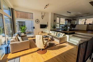 A spacious living area with a sofa, armchair, and a dining table. There is a TV mounted on the wall, a plant, and a staircase leading to another level.