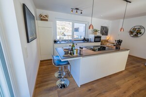A modern kitchen with a central island, bar stools, and a large window providing a view of the outdoors.