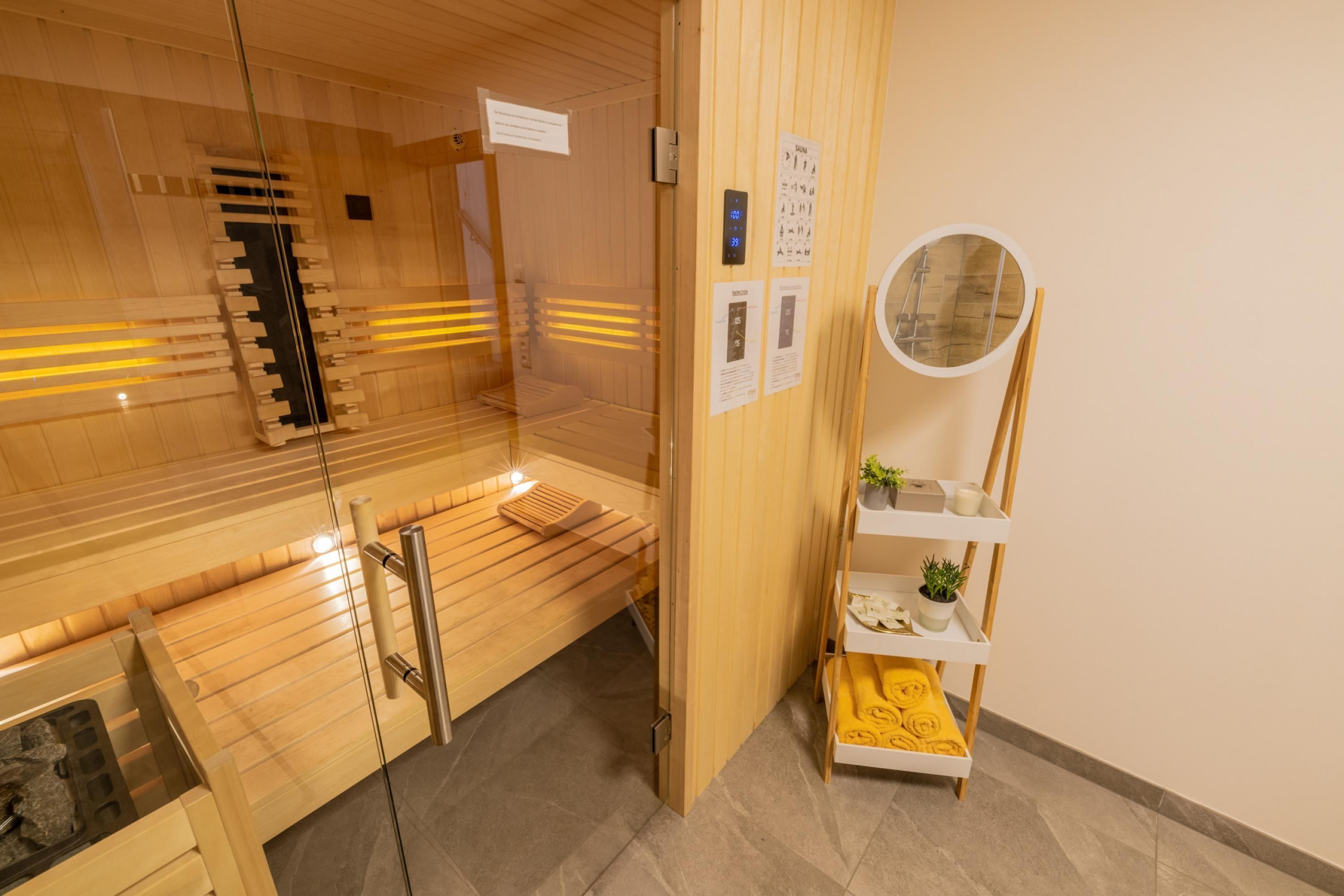 A sauna room with wooden benches, a glass door, a control panel, and a shelf with towels and a potted plant.
