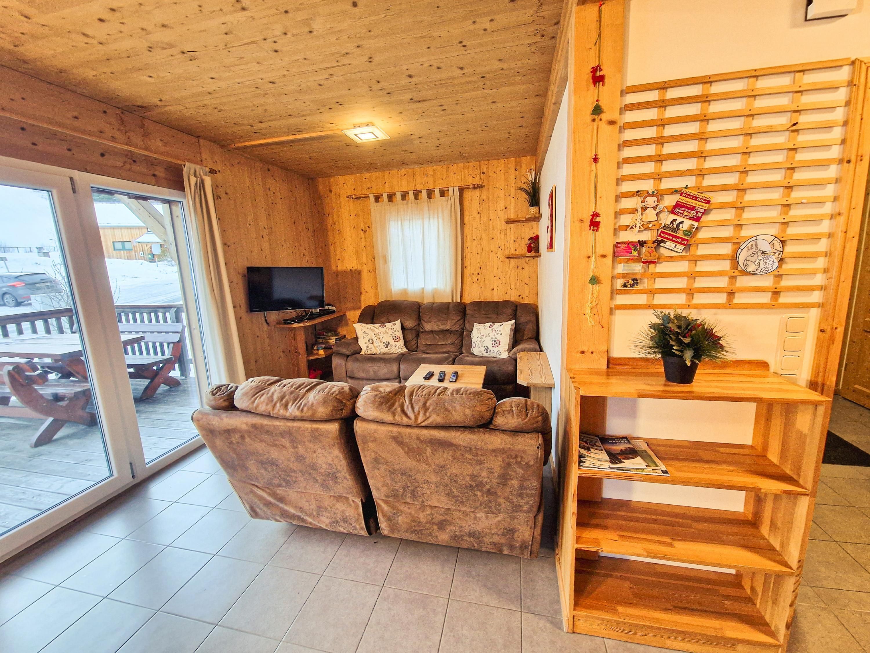 A cozy living room with a wooden ceiling, a sofa set, a TV, and a wooden shelf with decorations.
