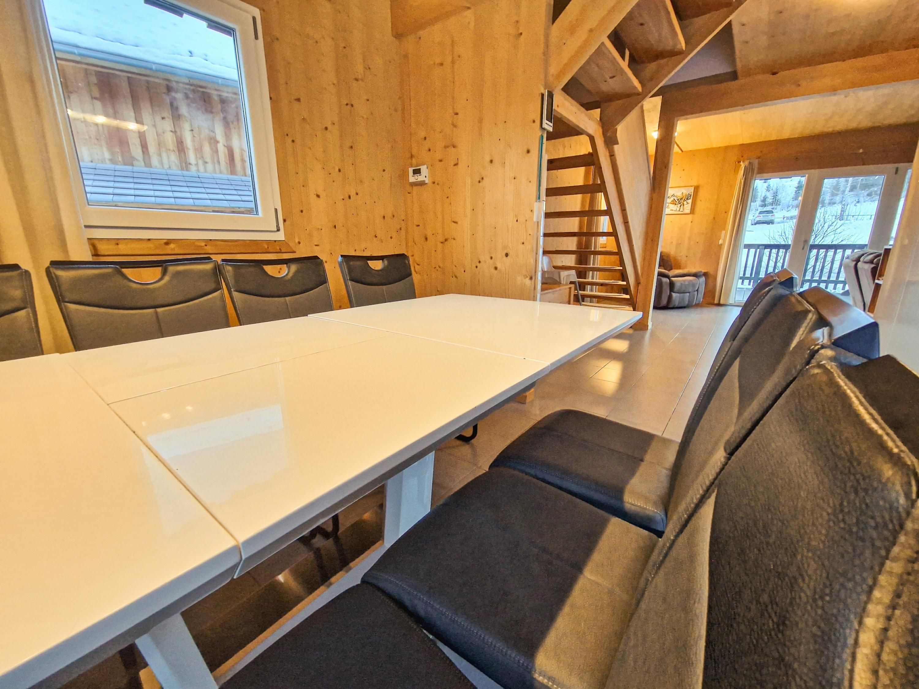 A wooden interior with a long table and six chairs, a staircase, and a view of the outside.