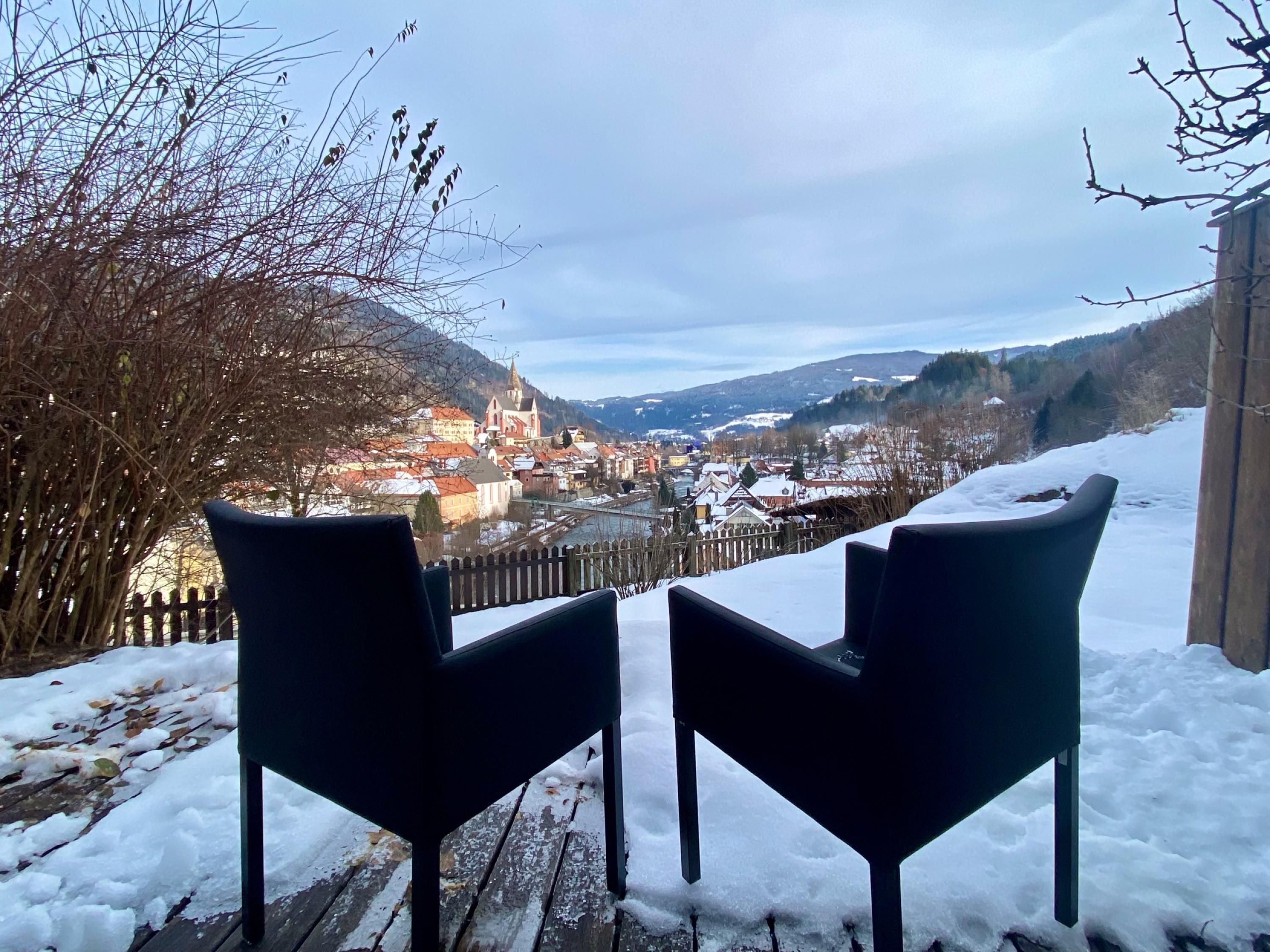 Dos sillas negras sobre una terraza de madera con nieve en el suelo, con vistas a un pueblo con edificios y un río.