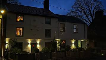 A two-story building with multiple windows, some of which are illuminated. There are outdoor lights along the front facade, and the building is surrounded by a stone wall and some greenery.