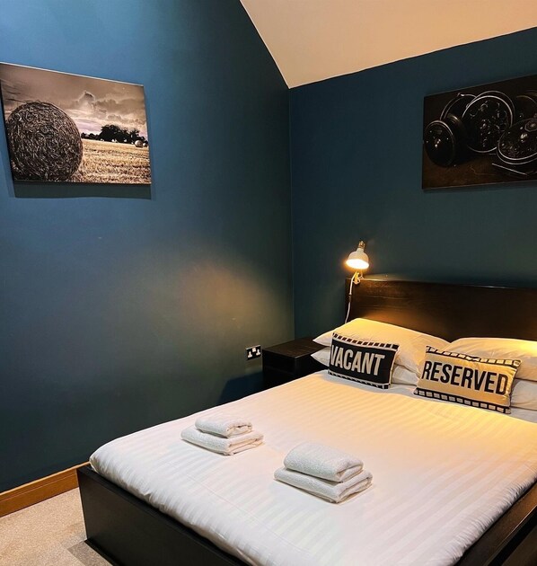 Un dormitorio con cama, dos almohadas, una mesita de noche, lámpara y un cuadro en la pared.
