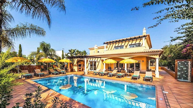 A pool area with lounge chairs and umbrellas, a pool with a tiled border, and a building with a tiled roof and windows. - Villa Cabrita de Parra (Albufeira)