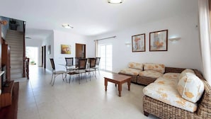 A spacious living room with a sofa, a dining table with chairs, a staircase, and a large window. - Villa Cordyline (Albufeira)