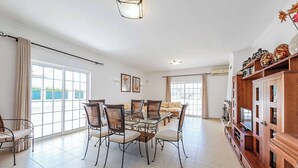 A spacious living room with a glass dining table surrounded by chairs, a sofa, and a wooden cabinet with decorative items. - Villa Cordyline (Albufeira)