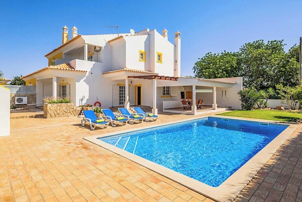 A two-story house with a swimming pool, a covered patio with lounge chairs, and a stone wall.