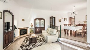 A living room with a fireplace, sofa, dining table, and staircase. - Villa Cardona (Albufeira)