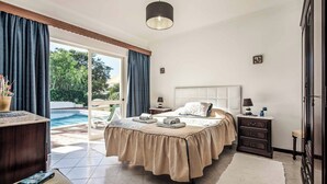 A bedroom with a bed, bedside tables, a nightstand, a desk, and a view of an outdoor swimming pool. - Villa Cardona (Albufeira)