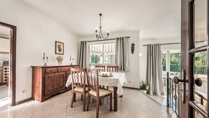 A dining area with a wooden table and chairs, a chandelier, and a view through a window with curtains. - Villa Cardona (Albufeira)