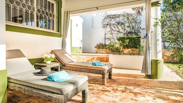 A sunlit outdoor lounge area with wicker furniture, a stone wall, and a view of the garden. - Villa Cardona (Albufeira)