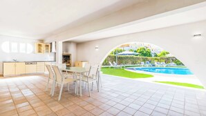 A modern kitchen with white cabinets and a dining area with a table and chairs. There is a view of a swimming pool and a garden with lounge chairs. - Villa Corcovada (Albufeira)