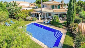 Une maison avec une piscine, entourée d’une végétation luxuriante, et un patio aménagé avec des chaises longues.