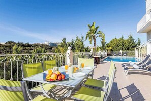 Un espace repas aménagé au bord de la piscine, avec une table dressée pour deux personnes, entourée de chaises vertes, et une piscine avec des chaises longues.