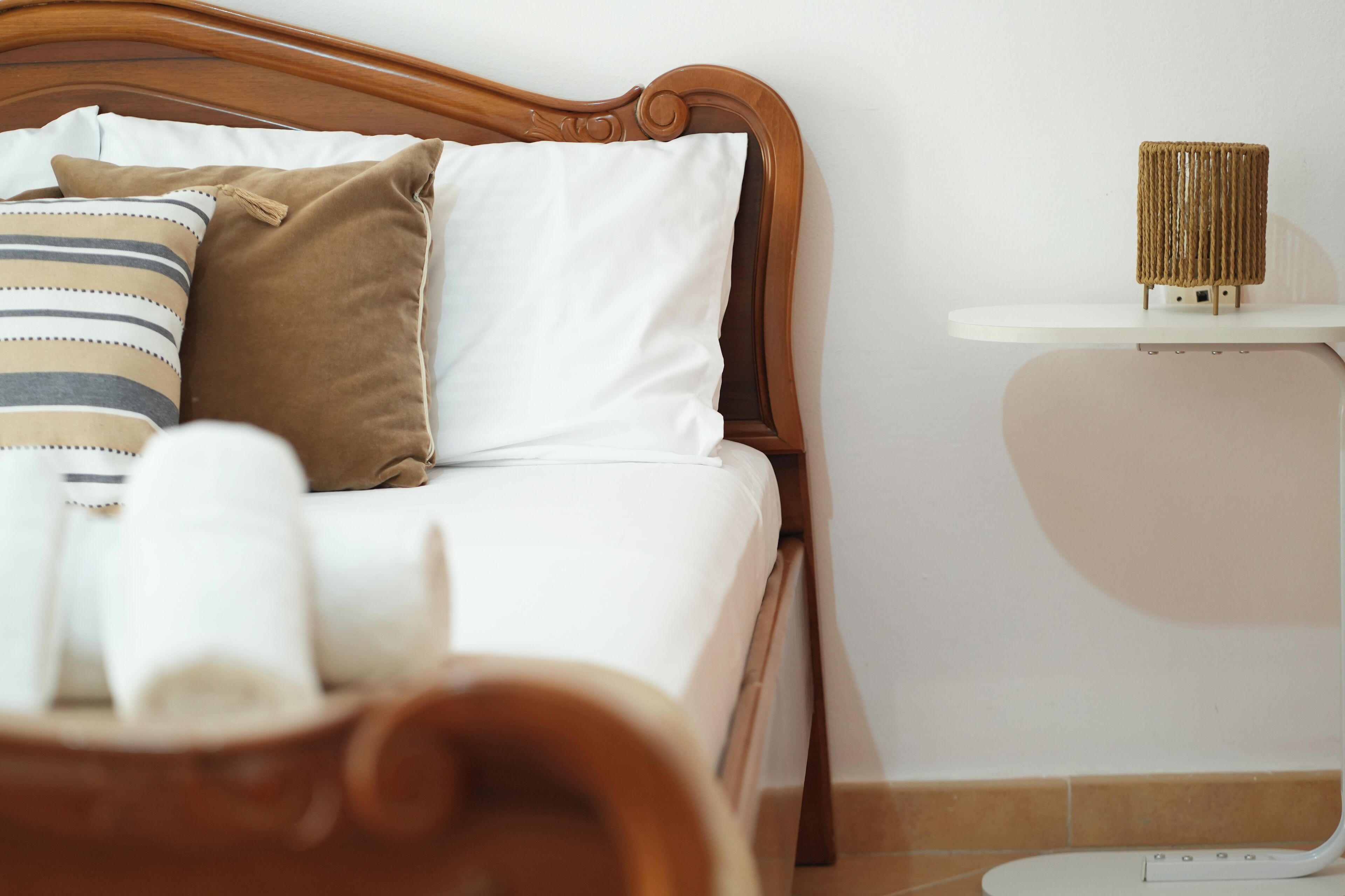 Una cama bien hecha con ropa de cama blanca y marrón, un cojín a rayas y una mesita de noche con una lámpara de mimbre.
