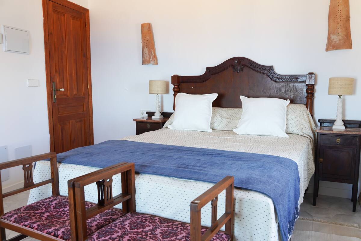 Quarto com cama de madeira, duas mesas de cabeceira com luminárias, uma cadeira de madeira e um banco estampado.