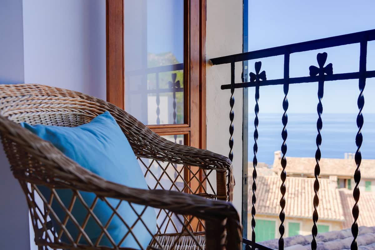 Uma cadeira de vime com almofada azul numa varanda com vista para edifícios e o mar.