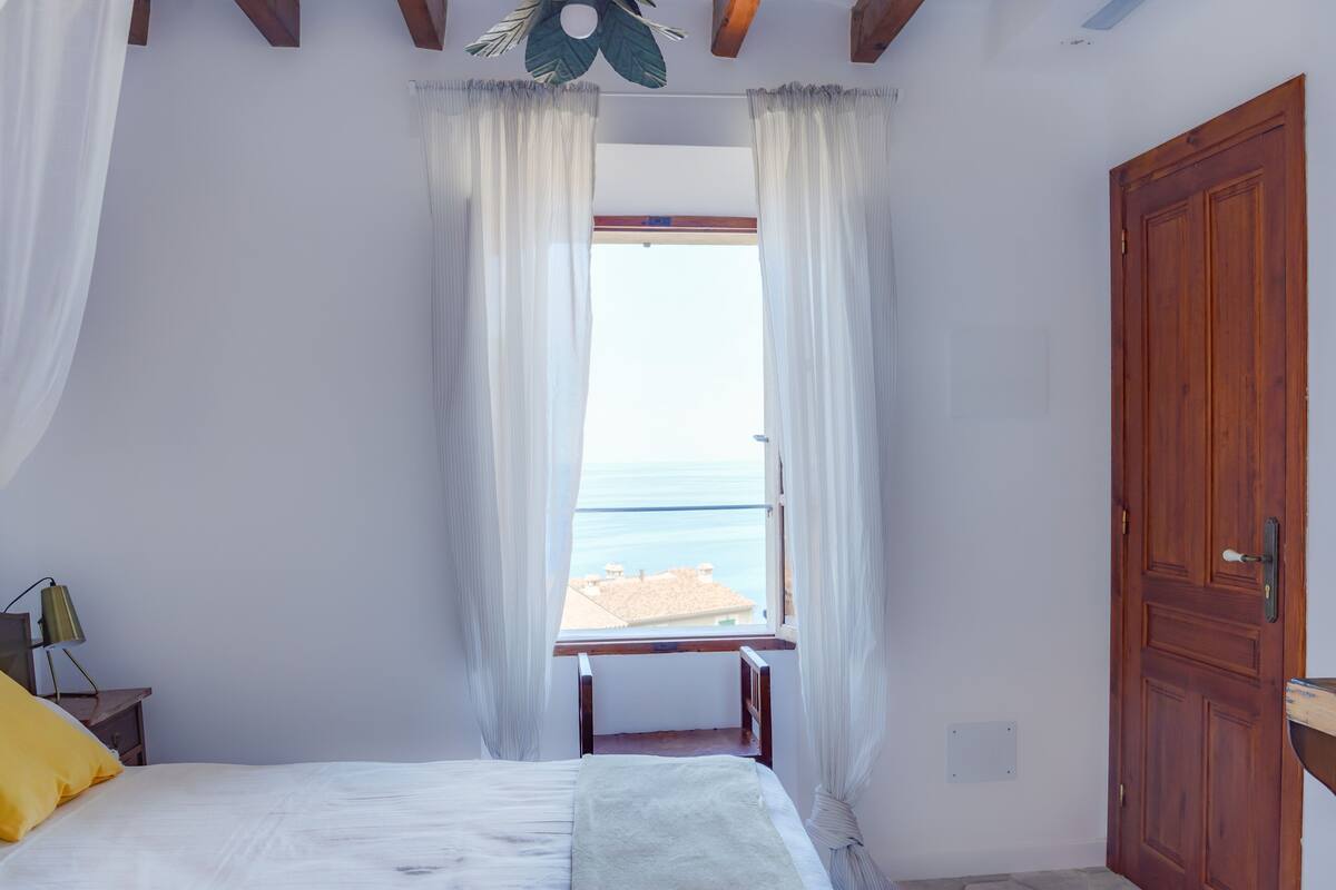 Quarto com cama, janela com cortinas, porta de madeira e vista para o mar.
