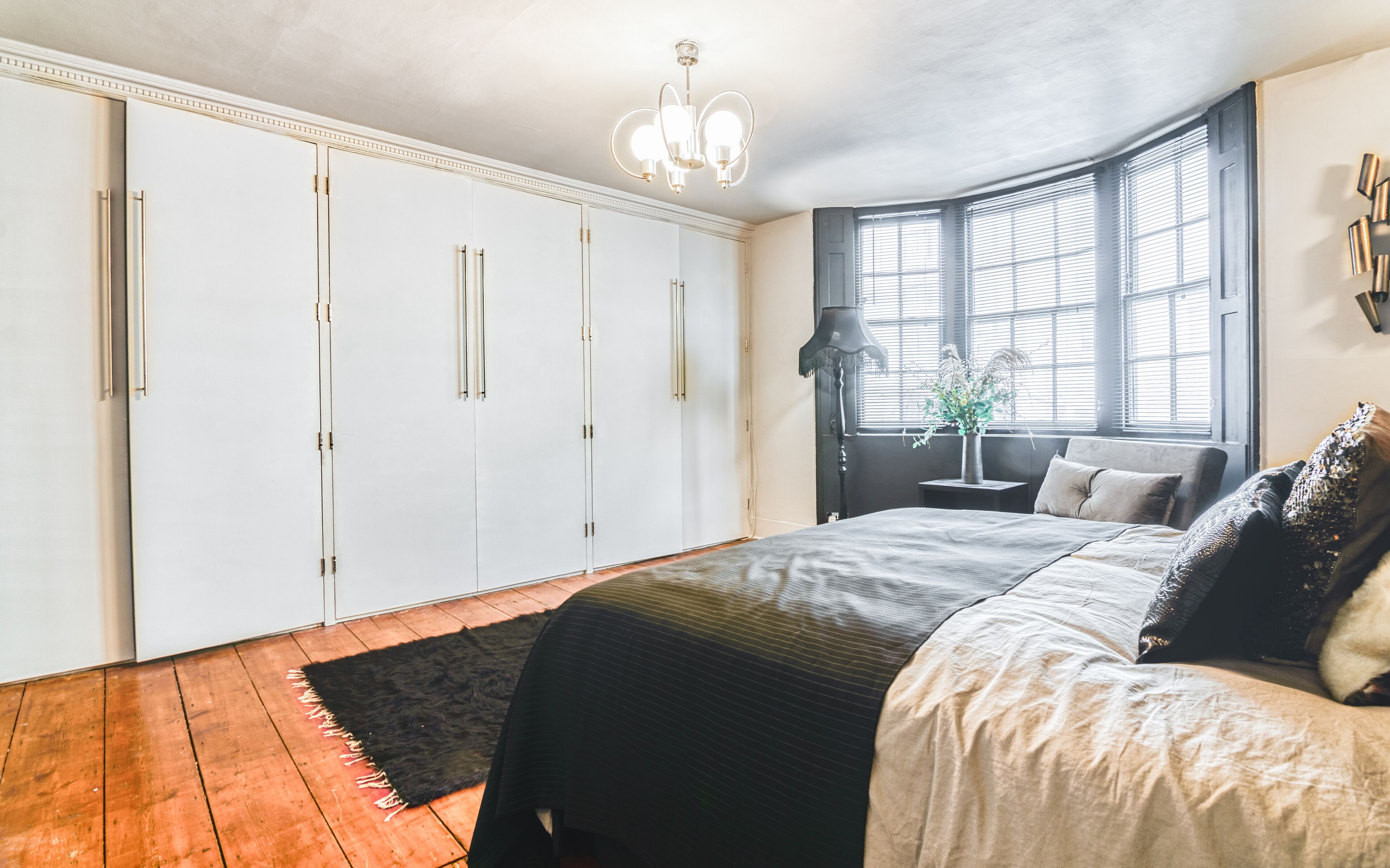 A bedroom with a large bed, a wardrobe, a desk, and a window with blinds.