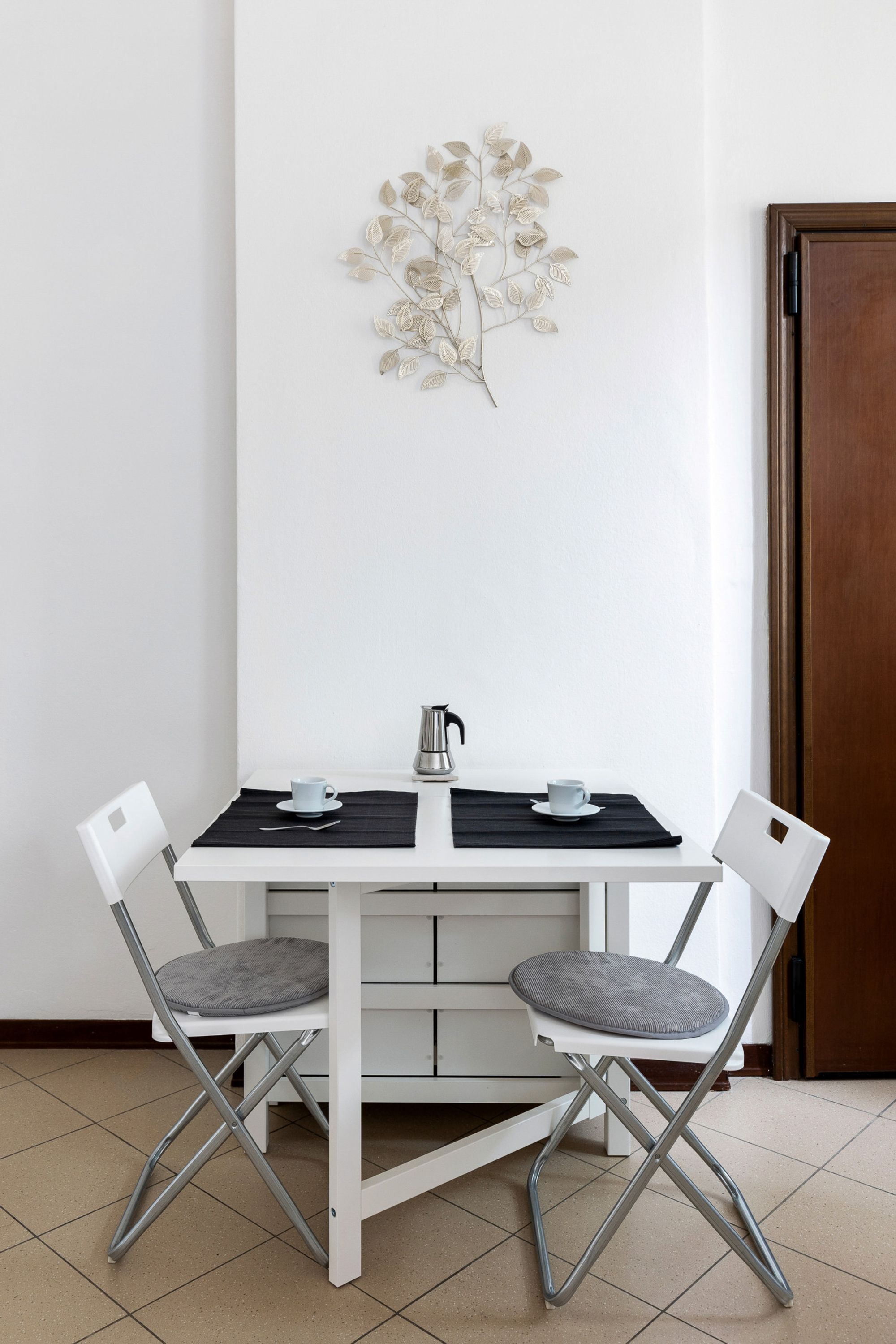 A small dining area with a white table, two chairs, a black placemat, a silver kettle, and a decorative wall piece.