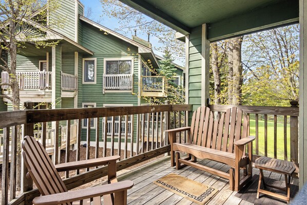 A wooden deck with Adirondack chairs, a wooden railing, and a view of a green house with multiple windows and balconies.