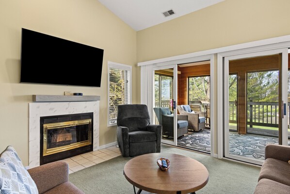 A living room with a fireplace, a flat-screen TV, a round coffee table, and a sliding glass door leading to an outdoor patio.