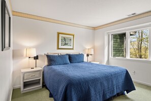 A bedroom with a large bed, two bedside lamps, a nightstand, a framed picture on the wall, and a window with a view of trees. - CrossWinds Getaway at Shanty Creek (bellaire)