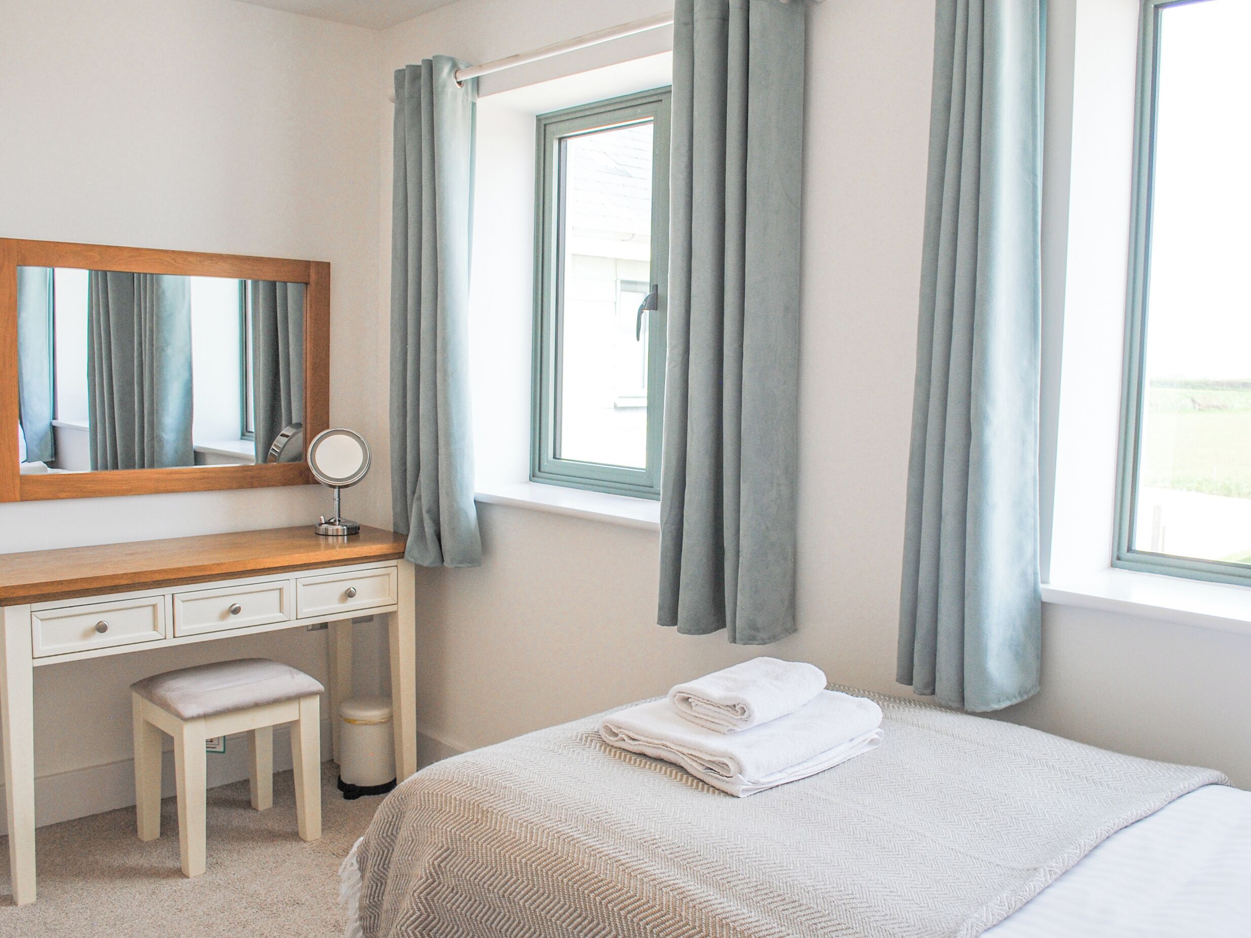 A bedroom with a bed, a vanity with a mirror, a small stool, and a window with curtains.