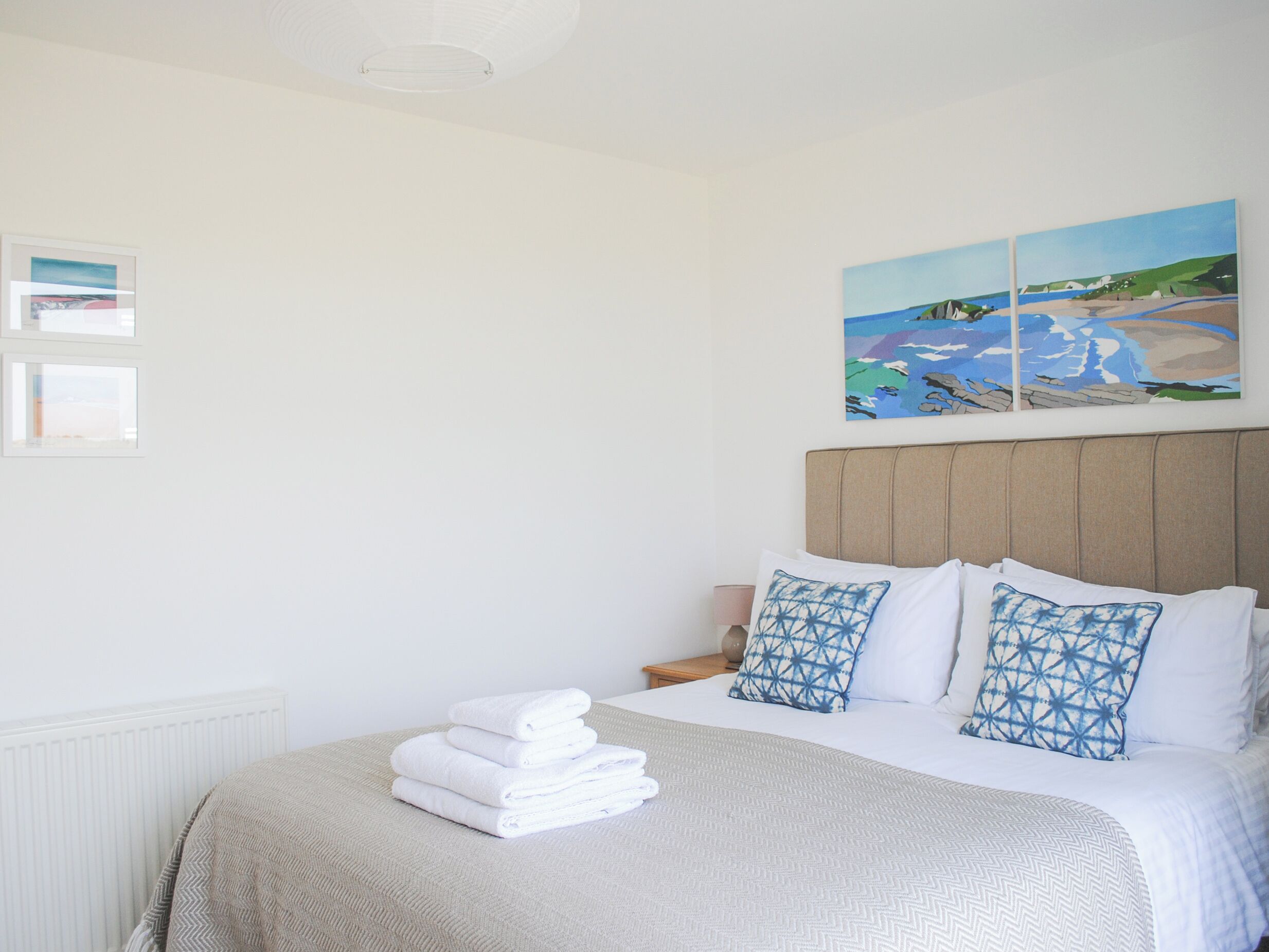 A neatly made bed with white linens and a patterned throw, a headboard with a light wood finish, and a painting of a coastal scene above the bed.
