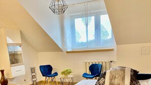 Un dormitorio con techo estilo loft, una cama grande con ropa de cama estampada, dos sillas azules, una mesita de noche con una planta, y una lámpara colgante.