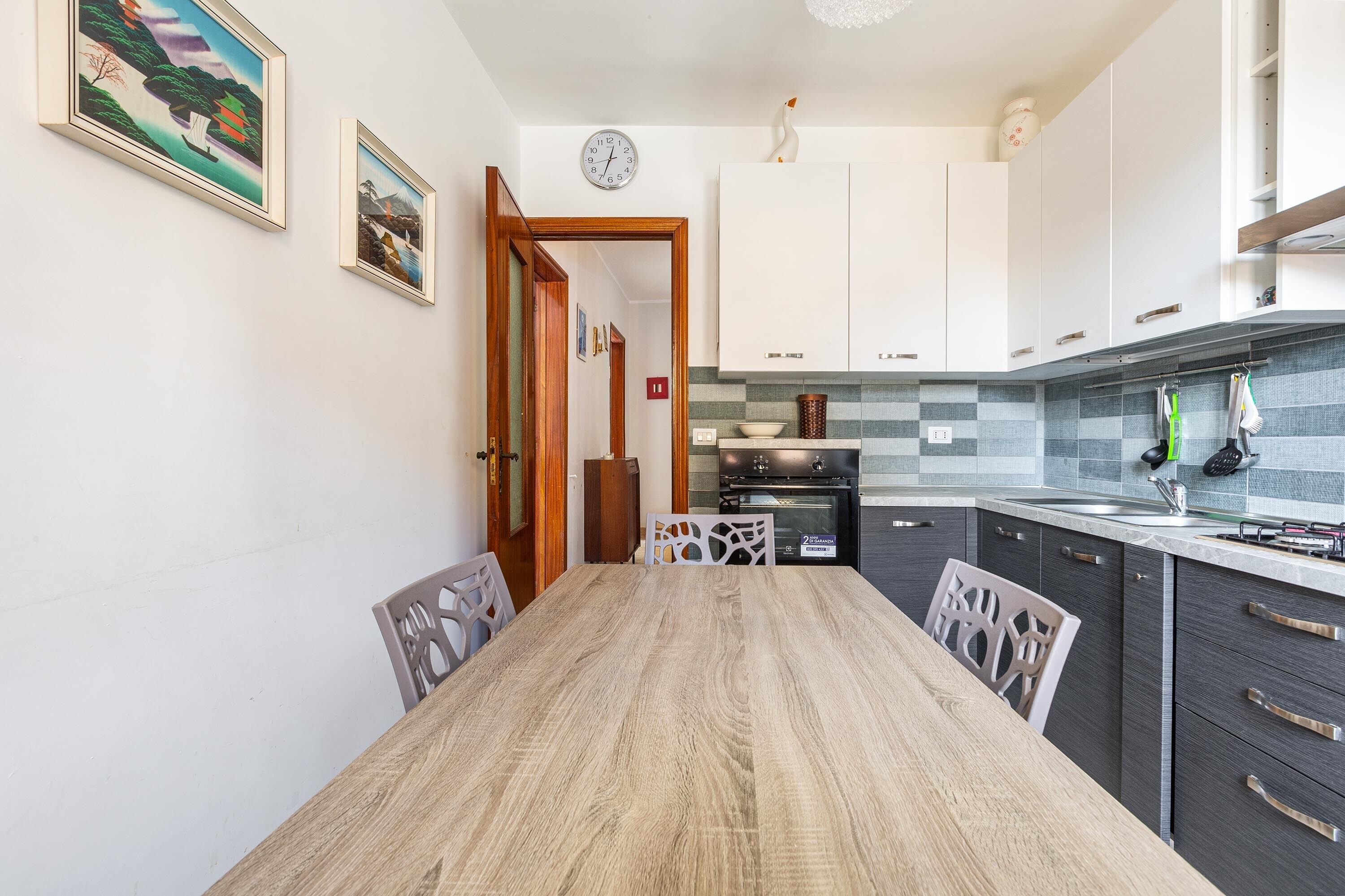 A modern kitchen with white cabinets, a wooden dining table, and a clock on the wall.