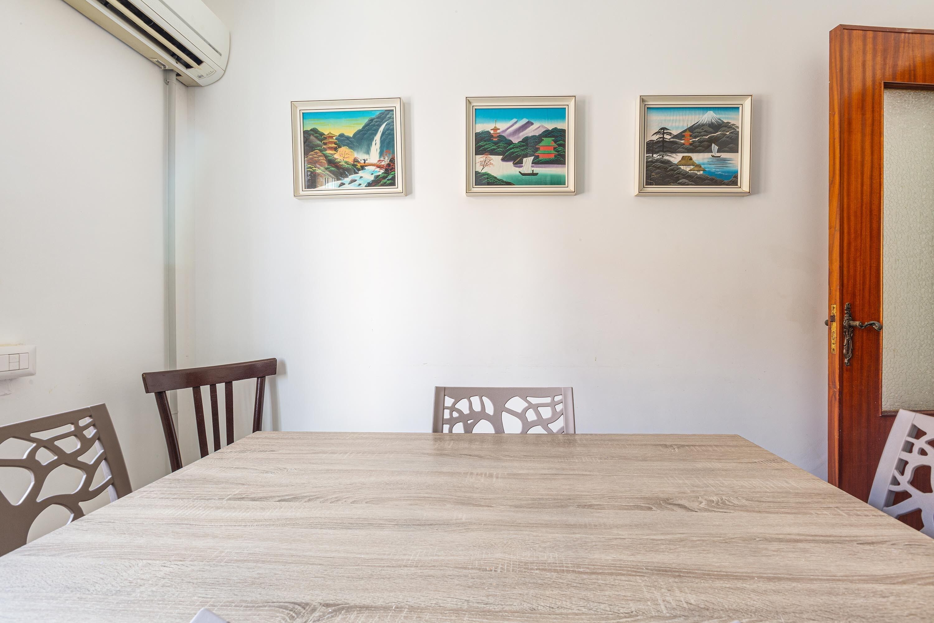 A dining area with a wooden table, two chairs, and three framed paintings on the wall.