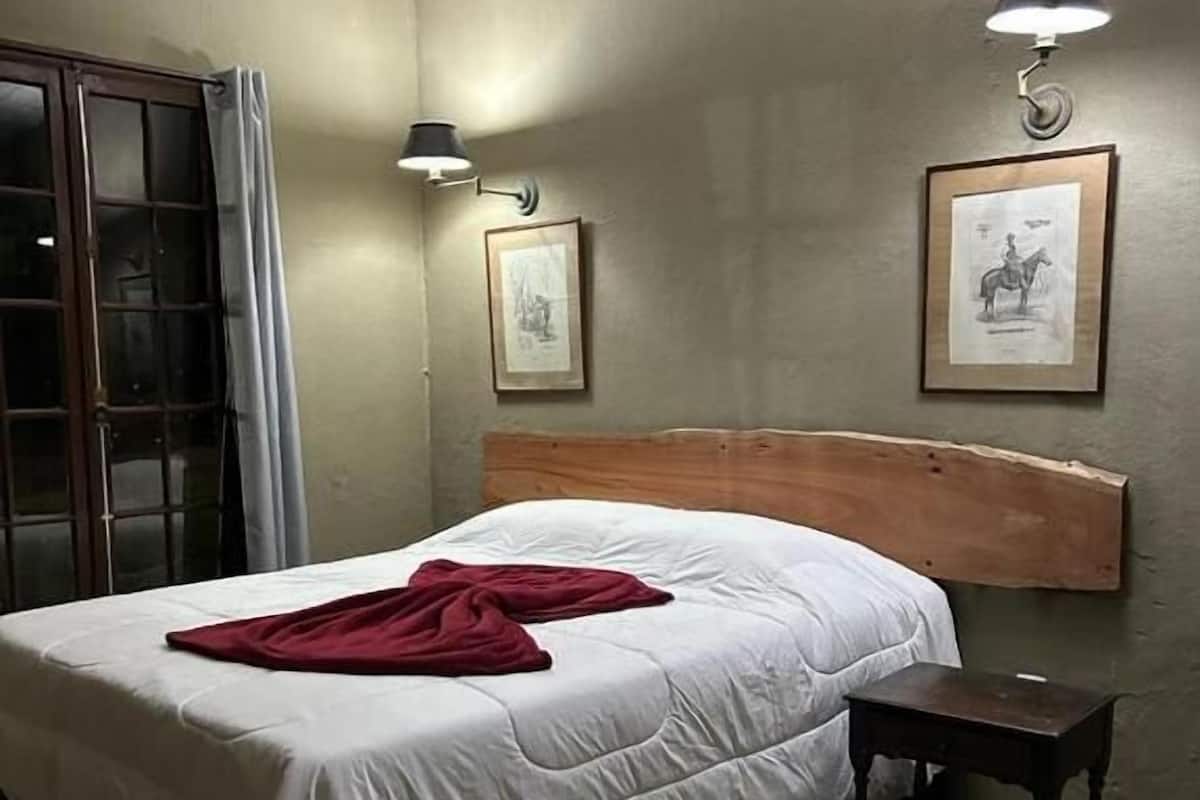 Quarto com cama, mesa de cabeceira, janela com cortinas e duas luminárias na parede.