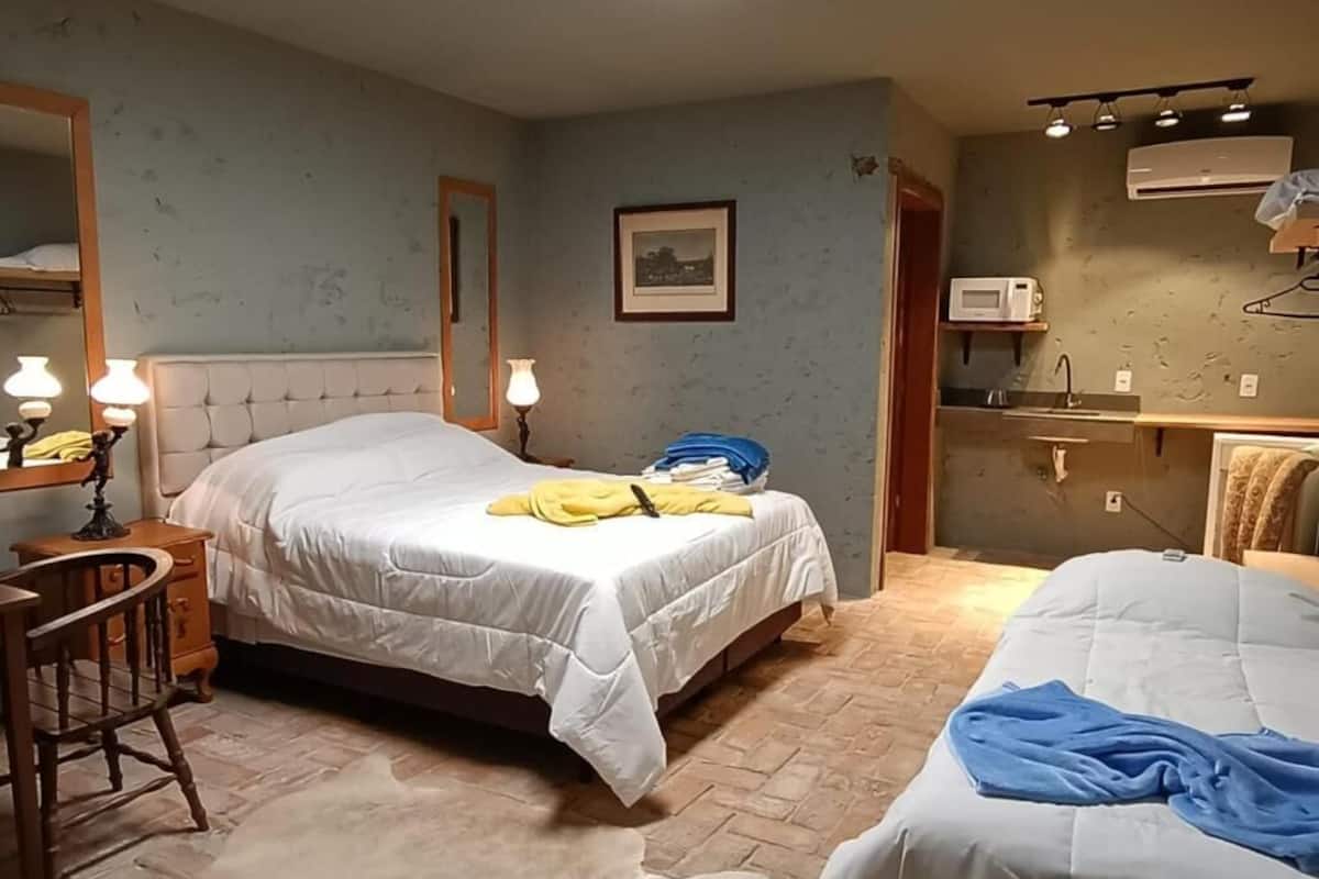 Quarto com duas camas, uma cadeira de madeira, mesa de cabeceira, espelho, quadro pendurado na parede e área de kitchenette com micro-ondas.