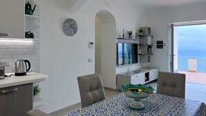 A modern kitchen with white cabinets, a kettle, and a toaster. There is a dining table with chairs and a decorative bowl. A television is mounted on the wall, and a window offers a view of the sea. - Villa Giulia lovely view #2 (Amalfi)