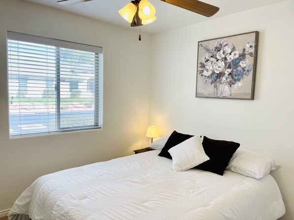 Une chambre à coucher avec un lit, une fenêtre avec des stores, un ventilateur de plafond, une lampe fixée au mur et un tableau représentant des motifs floraux.