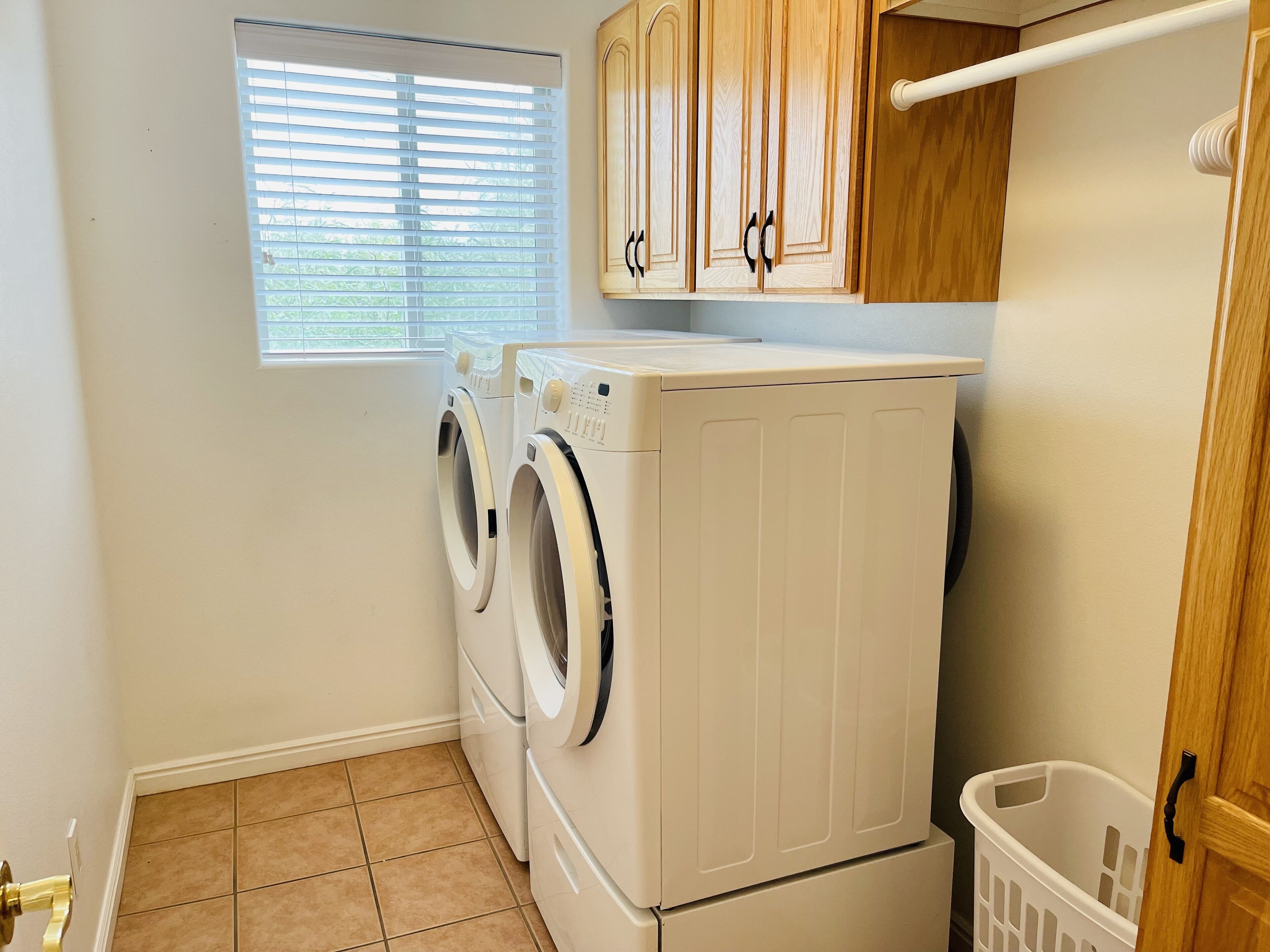 A laundry room with two front-loading washers and dryers, a window with blinds, wooden cabinets, and a white laundry basket.