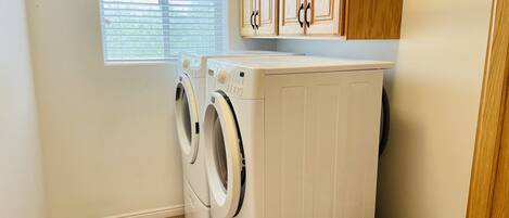 A laundry room with two front-loading washers and dryers, a window with blinds, wooden cabinets, and a white laundry basket.