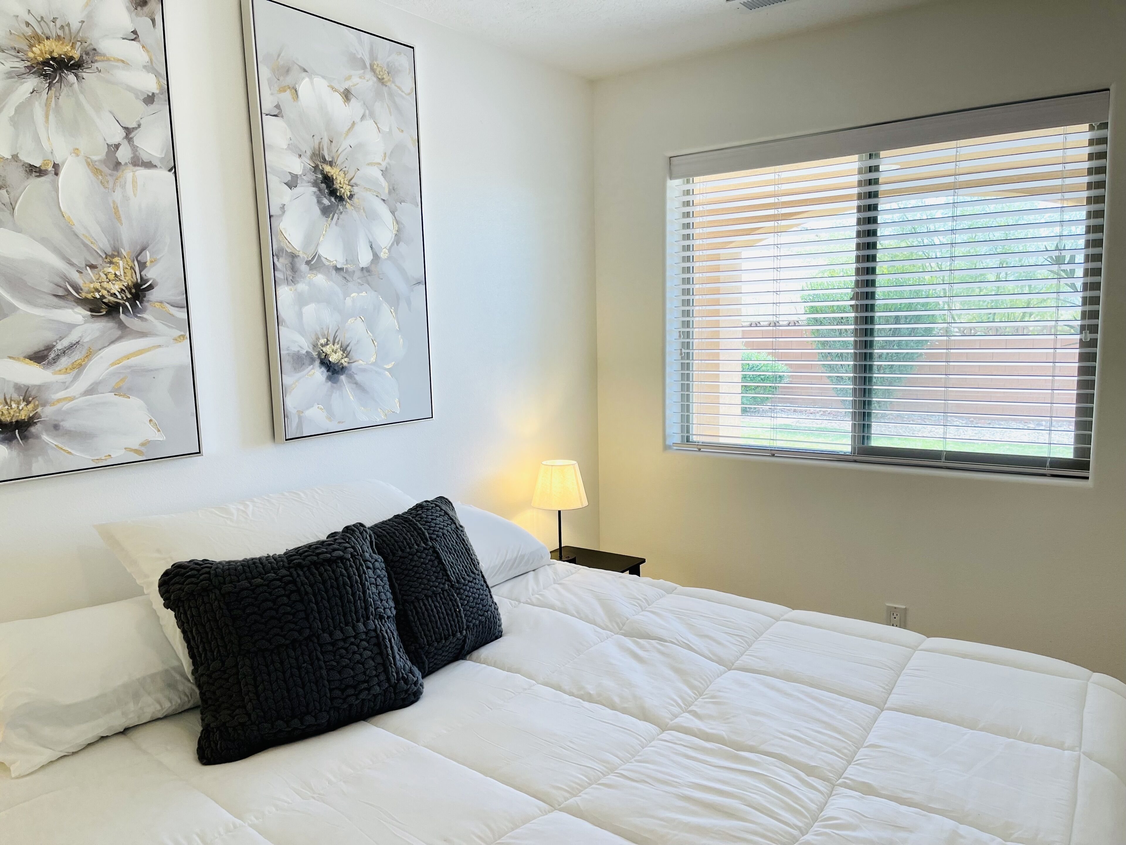 A bedroom with a large bed, white bedding, two black pillows, a bedside lamp, and two floral paintings on the wall.