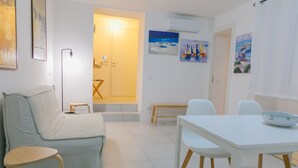 A modern, well-lit room with a sofa, a dining table with chairs, a small stool, and a door leading to another room. - A due Passi dal Molo by Wonderful Italy - Apartment (Cefalù)