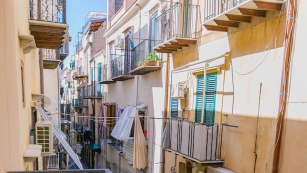 A narrow street with buildings on either side, balconies, and a blue car parked on the left. - A due Passi dal Molo by Wonderful Italy - Apartment (Cefalù)