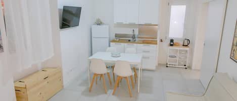A compact kitchen with white cabinets, a dining table with chairs, a TV mounted on the wall, and a window with curtains.