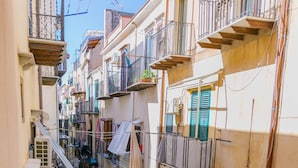 A narrow street with buildings on either side, balconies with railings, and a blue car parked on the left.